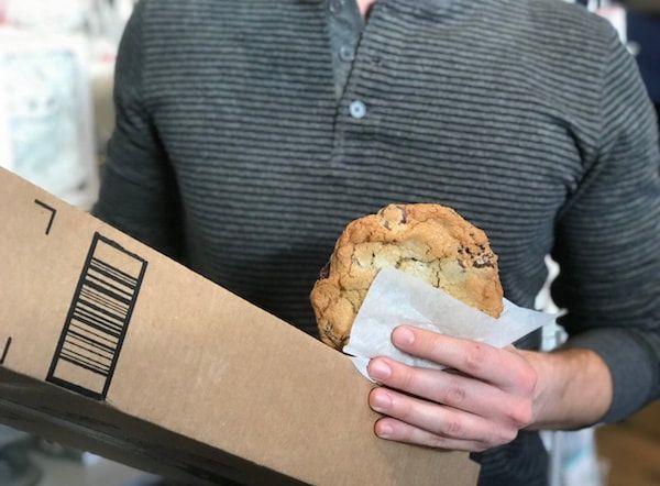Person holding a large cookie and an Amazon box in a home setting
