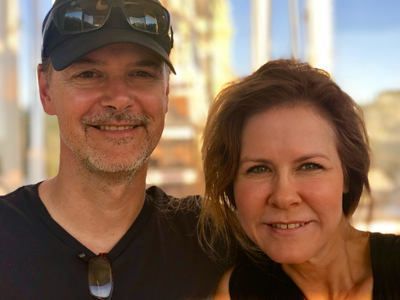 Smiling couple posing outdoors in casual clothes, with sunlit buildings in the background.