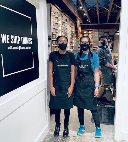 Two masked staff in aprons stand in a warehouse hallway beside a “WE SHIP THINGS” sign.