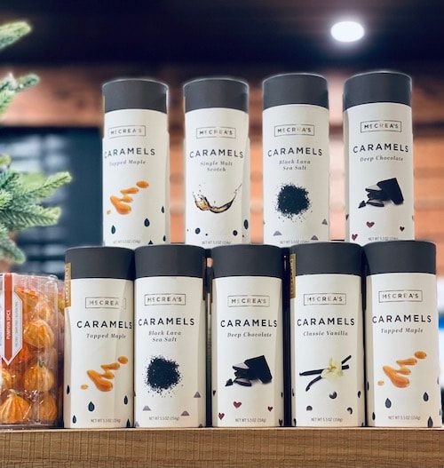 Stacked Caramels dessert jars in white and black packaging, displayed on wooden shelves with plant decor.