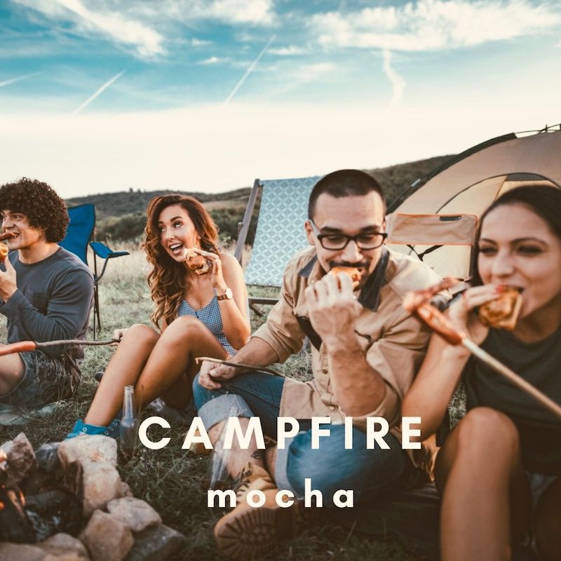 Friends sitting around a campfire at sunset, roasting food with tents in the background.