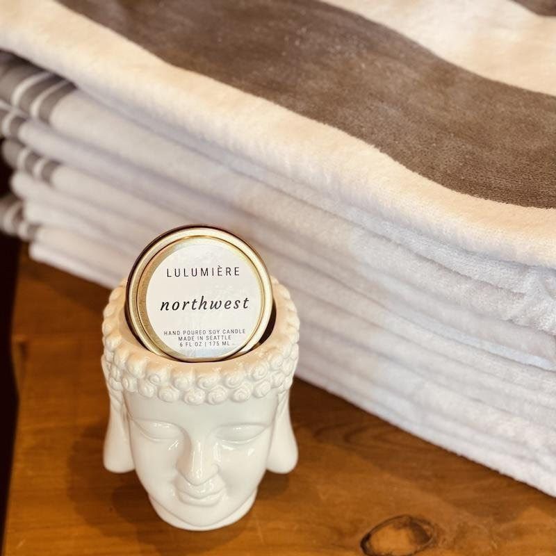 White Buddha-head candle with round lid beside folded beige and brown blanket on a wooden surface