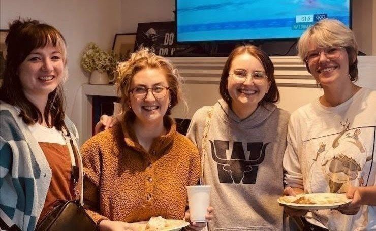 Four friends smiling indoors, standing together with drinks and a plate of food