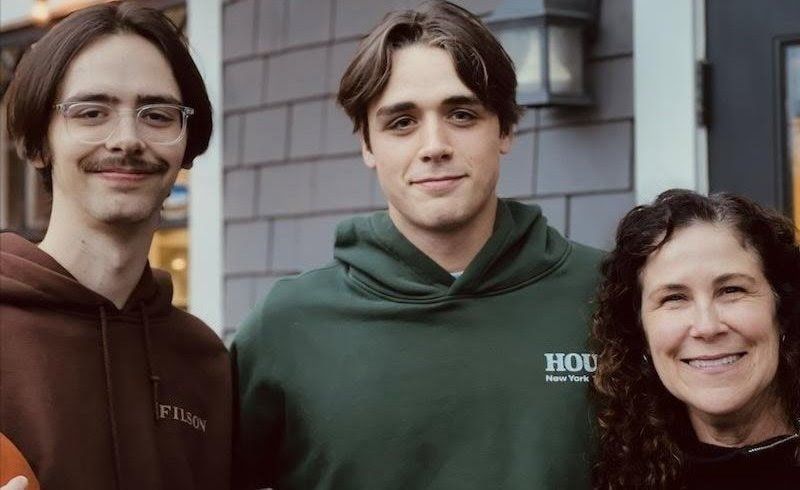 Three people standing outdoors, smiling at the camera, wearing brown and green hoodies.