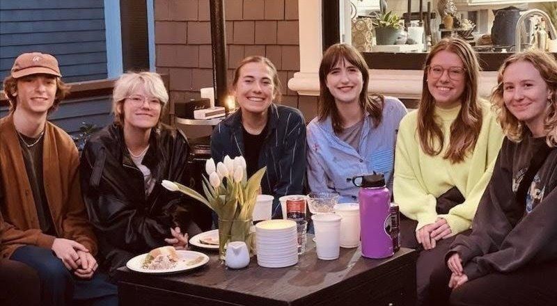 Six friends smiling at an outdoor cafe table with drinks and plates, posing together
