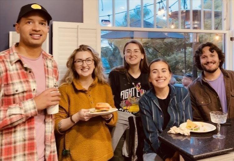 Five people smiling and holding plates at a casual indoor gathering near large windows