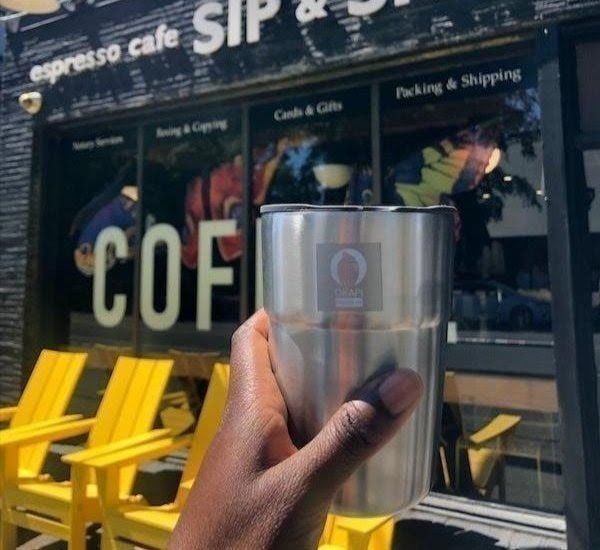 Hand holding a stainless steel cup in front of a coffee shop with bright yellow chairs