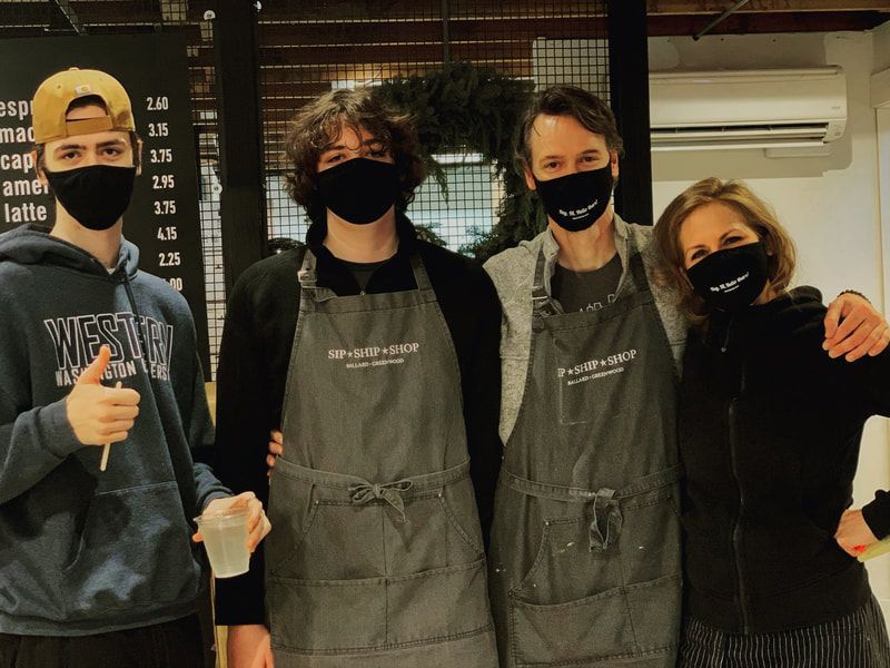 Four masked café staff in aprons posing together indoors, one giving a thumbs-up.