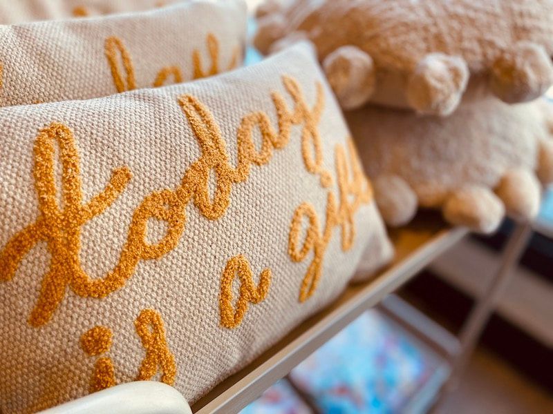 Embroidered cream throw pillow with orange script, beside a matching tasseled pillow on a shelf