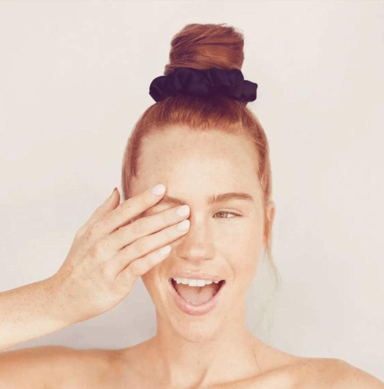 Woman with a top bun covering one eye, smiling against a plain background