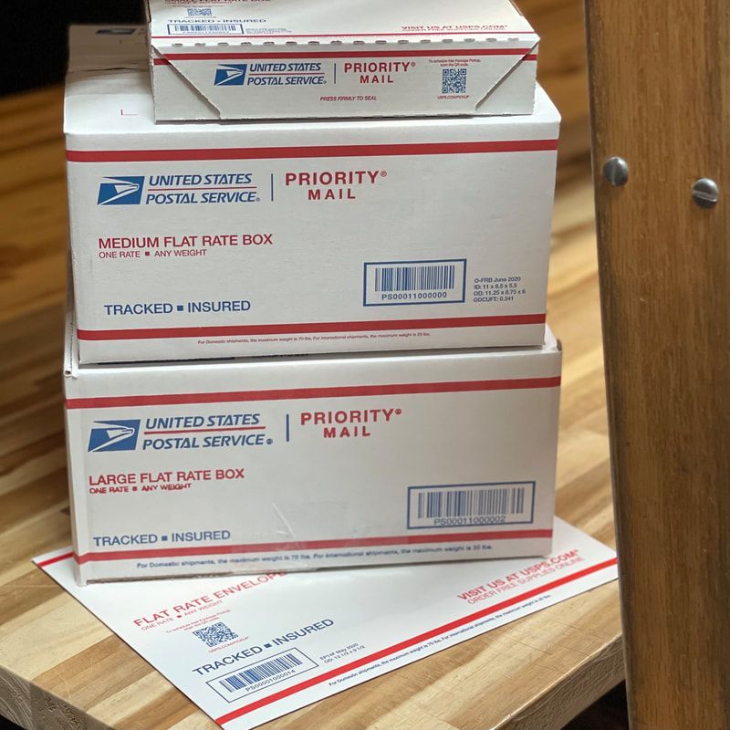 Stacked USPS Priority Mail boxes on a wooden table, with one flat-rate box lying in front.