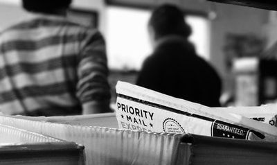 A stack of mail with a Priority Mail envelope in the foreground and two blurred people in the background.