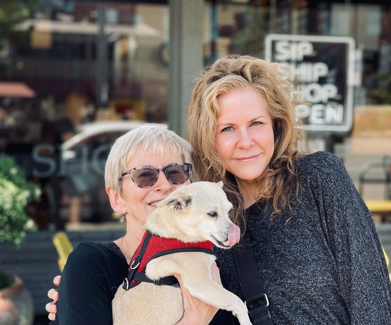 Two women holding a small white dog in front of a storefront window with an “OPEN” sign.