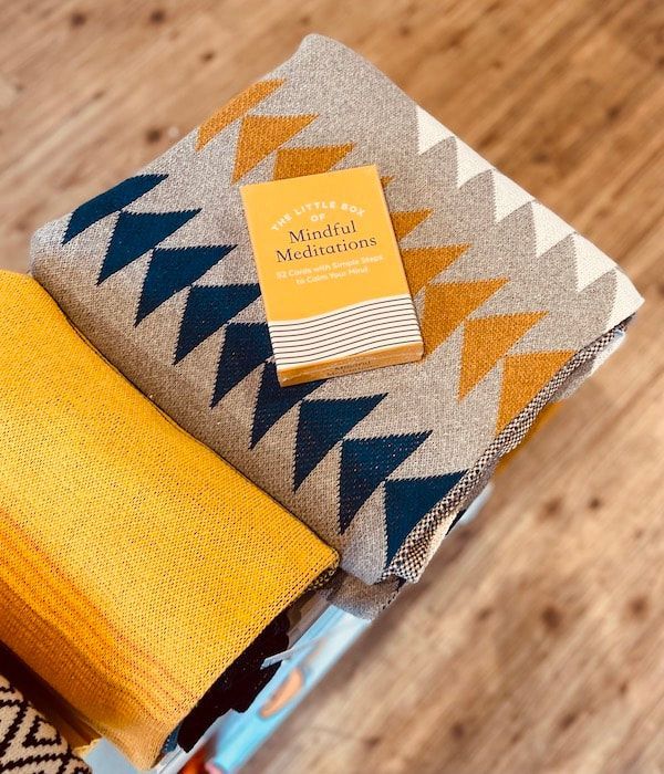 Folded patterned blanket in beige, navy, and mustard with a small yellow-and-white striped booklet on top.