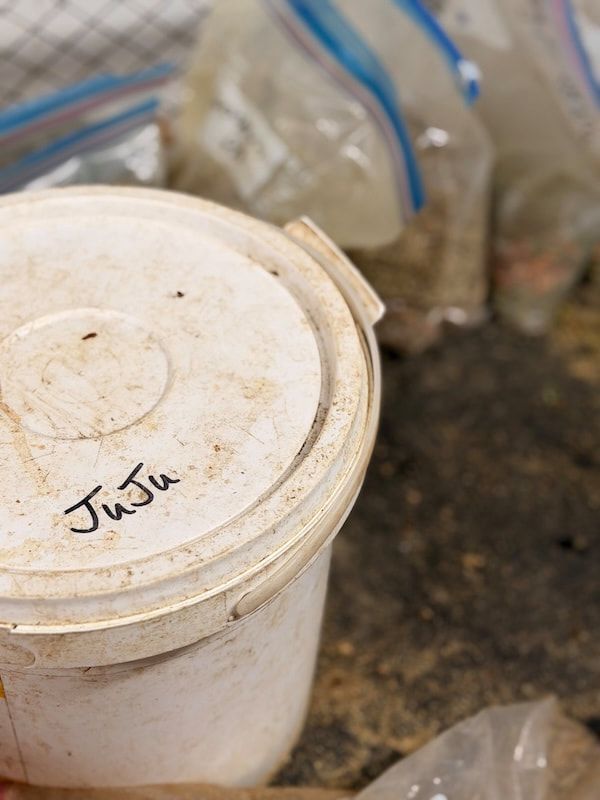 White plastic bucket with a handwritten label on dusty ground beside a clear plastic bag.