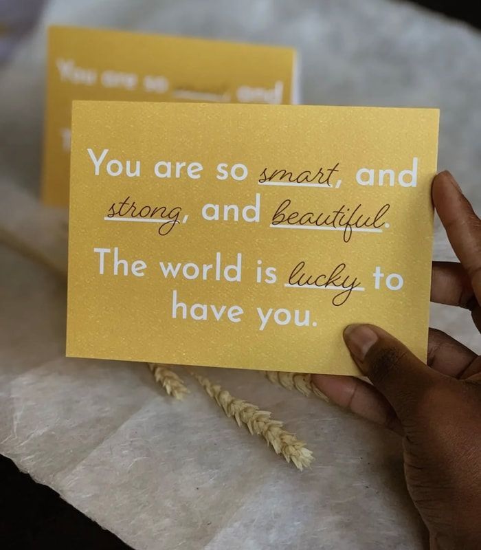Hand holding a yellow note with a handwritten uplifting message, with another blurred note behind it.