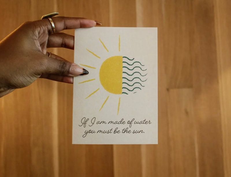 Hand holding a white card with a yellow sun and wavy lines over a quote on a wood background