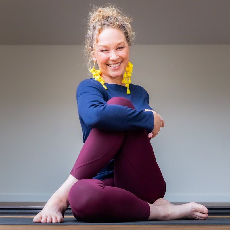 Smiling person sitting cross-legged on a yoga mat indoors, wearing blue and maroon workout clothes.