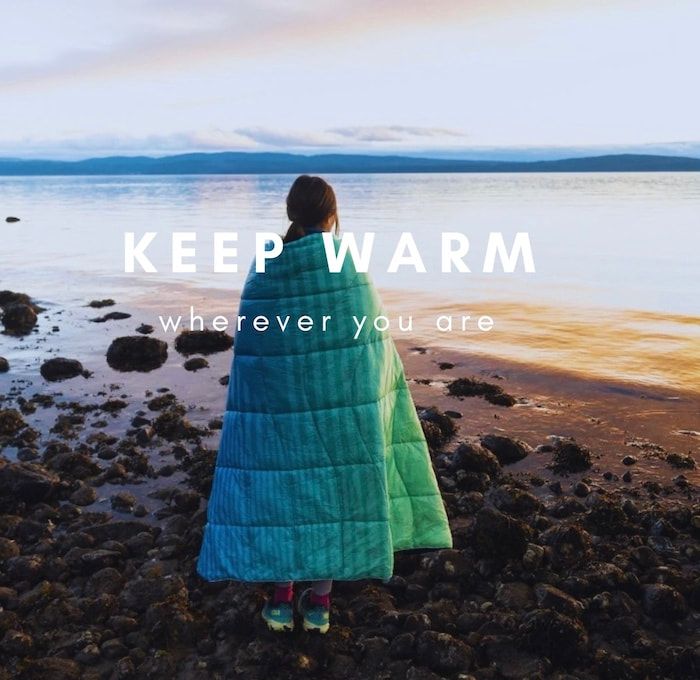 Person wrapped in a teal blanket stands on a rocky shore by a calm lake with “KEEP WARM wherever you are” text.