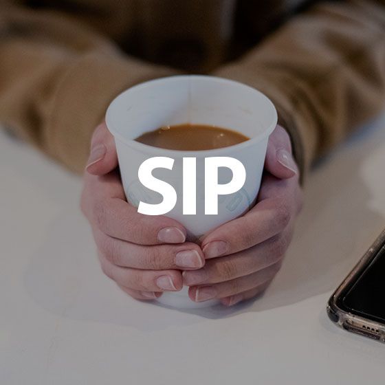 Hands holding a white cup of coffee on a table, with the word “SIP” overlaid.