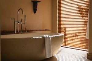Modern bathroom with a freestanding tub, chrome faucet, and sunlight through wooden blinds.