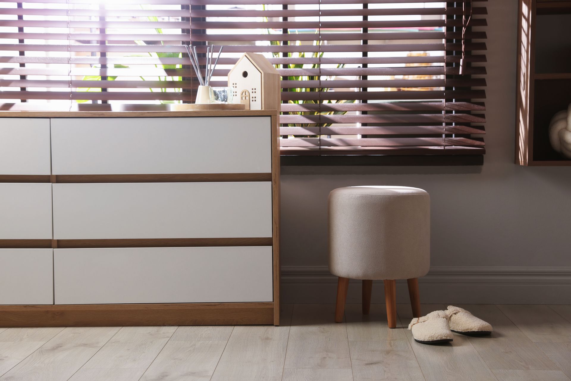 Modern bedroom with white dresser, window blinds, gray ottoman, and slippers on the floor Modern bedroom with white dresser, window blinds, gray ottoman, and slippers on the floor
