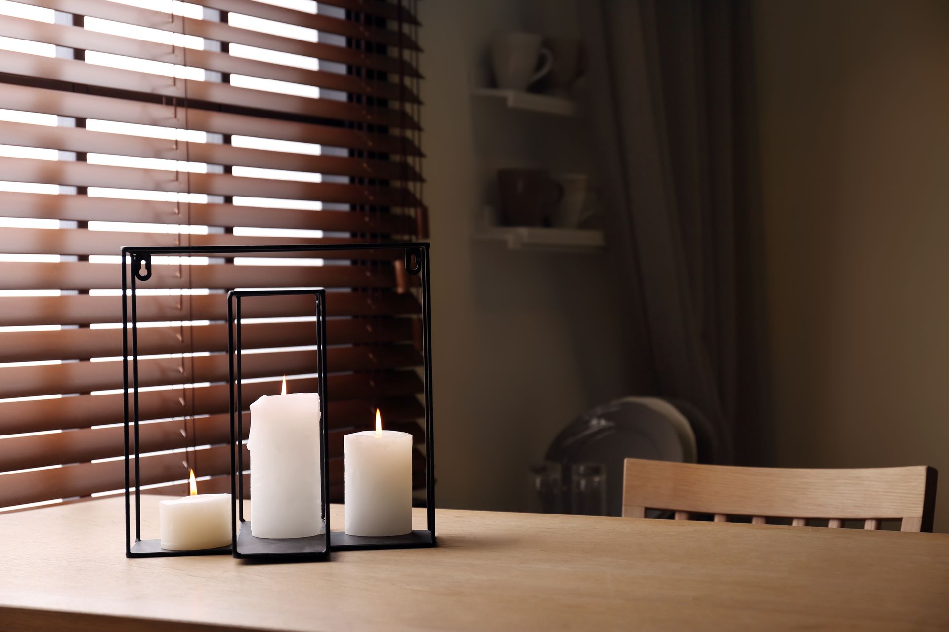 Candles on a table beside a window with wooden blinds in a softly lit room