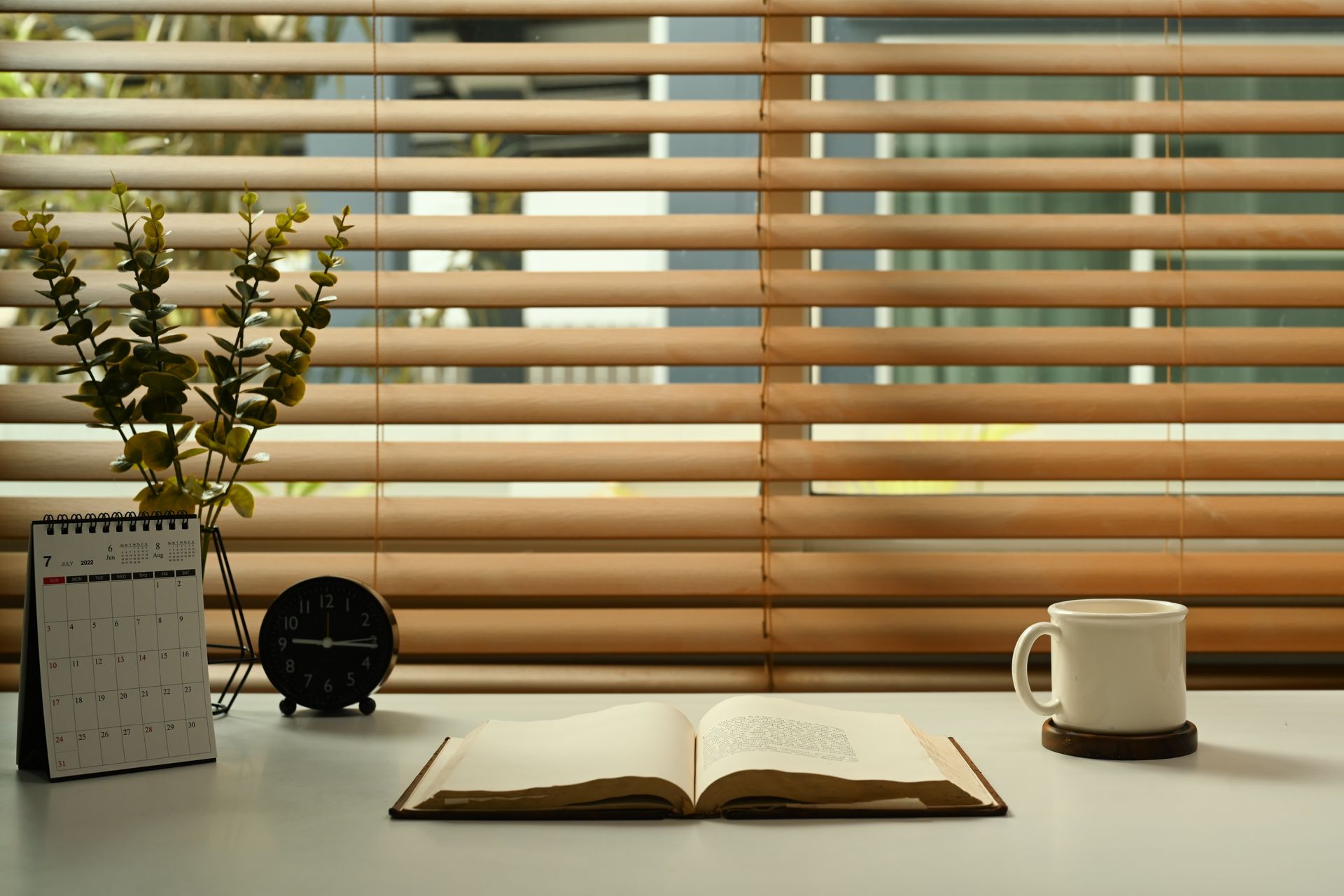 Open book on a desk beside a coffee mug, calendar, flowers, and a clock in front of window blinds
