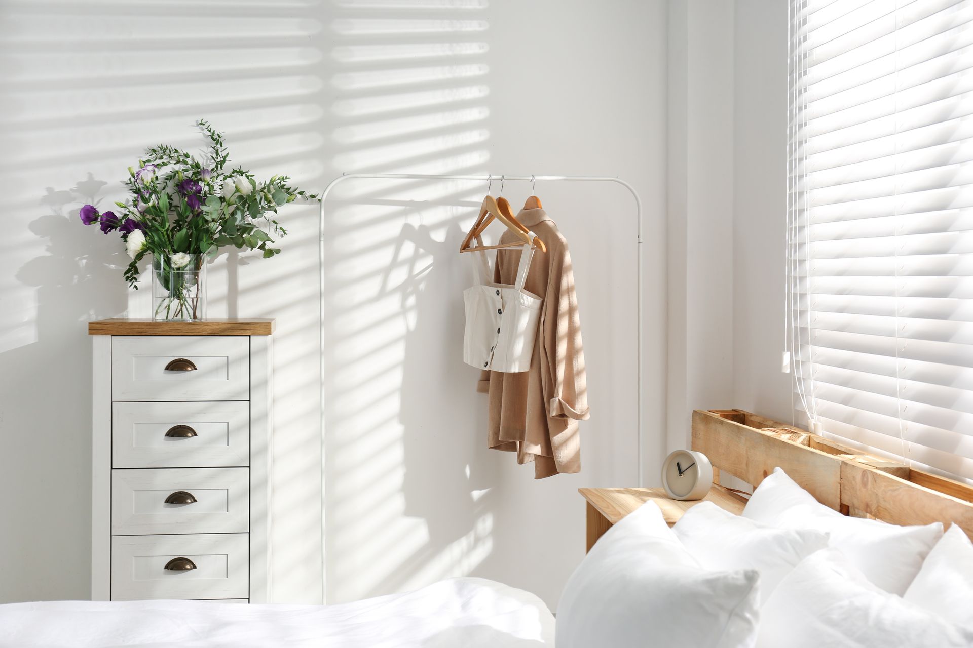 Sunlit bedroom with white dresser, flowers, hanging clothes, and bed by window blinds