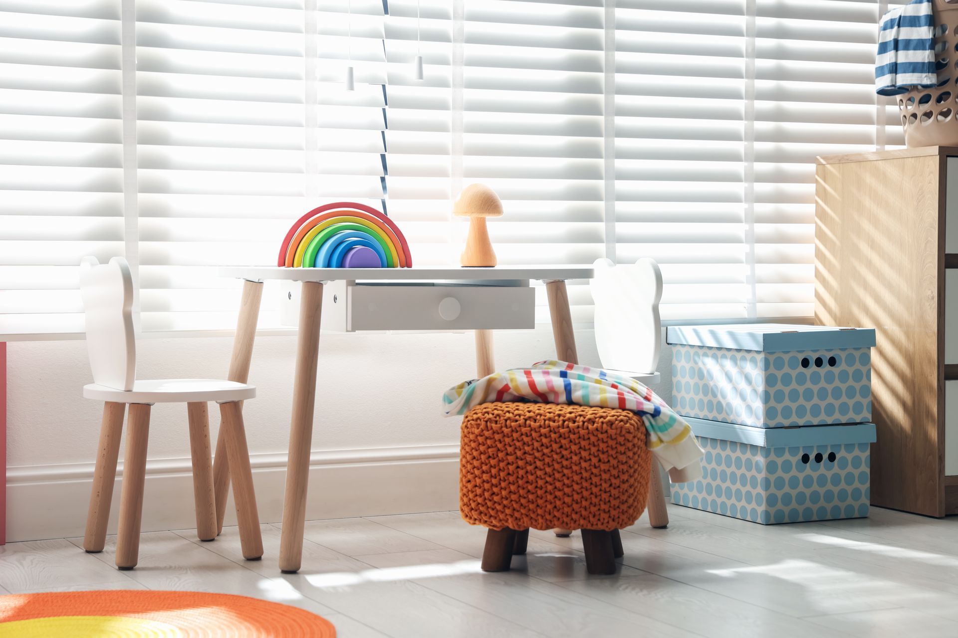 Bright children’s playroom with white desk, rainbow decor, toy bin, and blinds by a window Bright children’s playroom with white desk, rainbow decor, toy bin, and blinds by a window