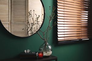 Round mirror above a dark table with branches in a vase, red candle, and window blinds in a green room