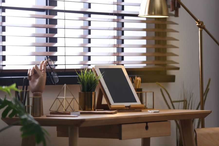 Desk with tablet, lamp, plant, and framed art by a window with blinds.