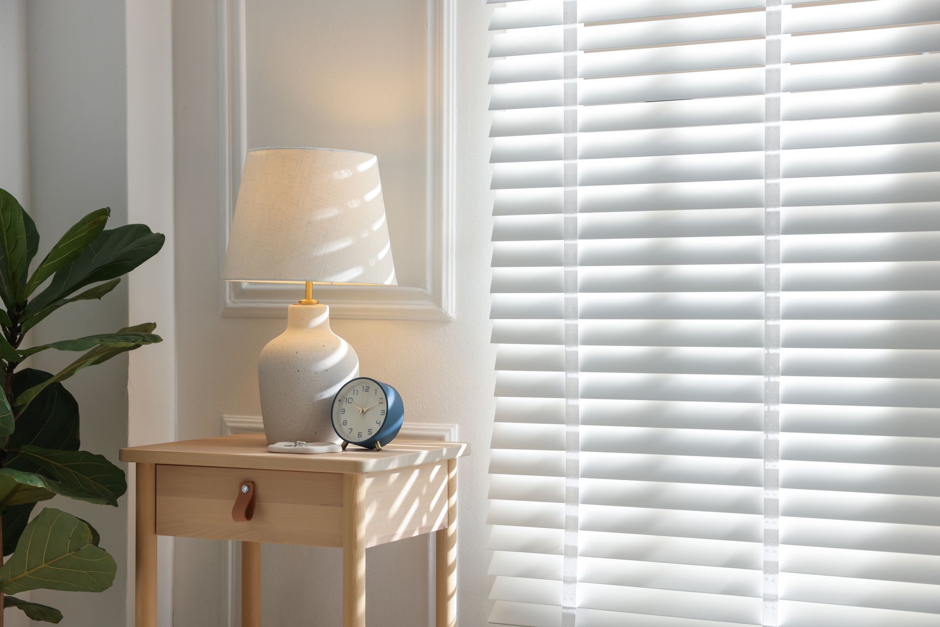 Sunlit room with white blinds, side table, lamp, clock, and a leafy plant by the window