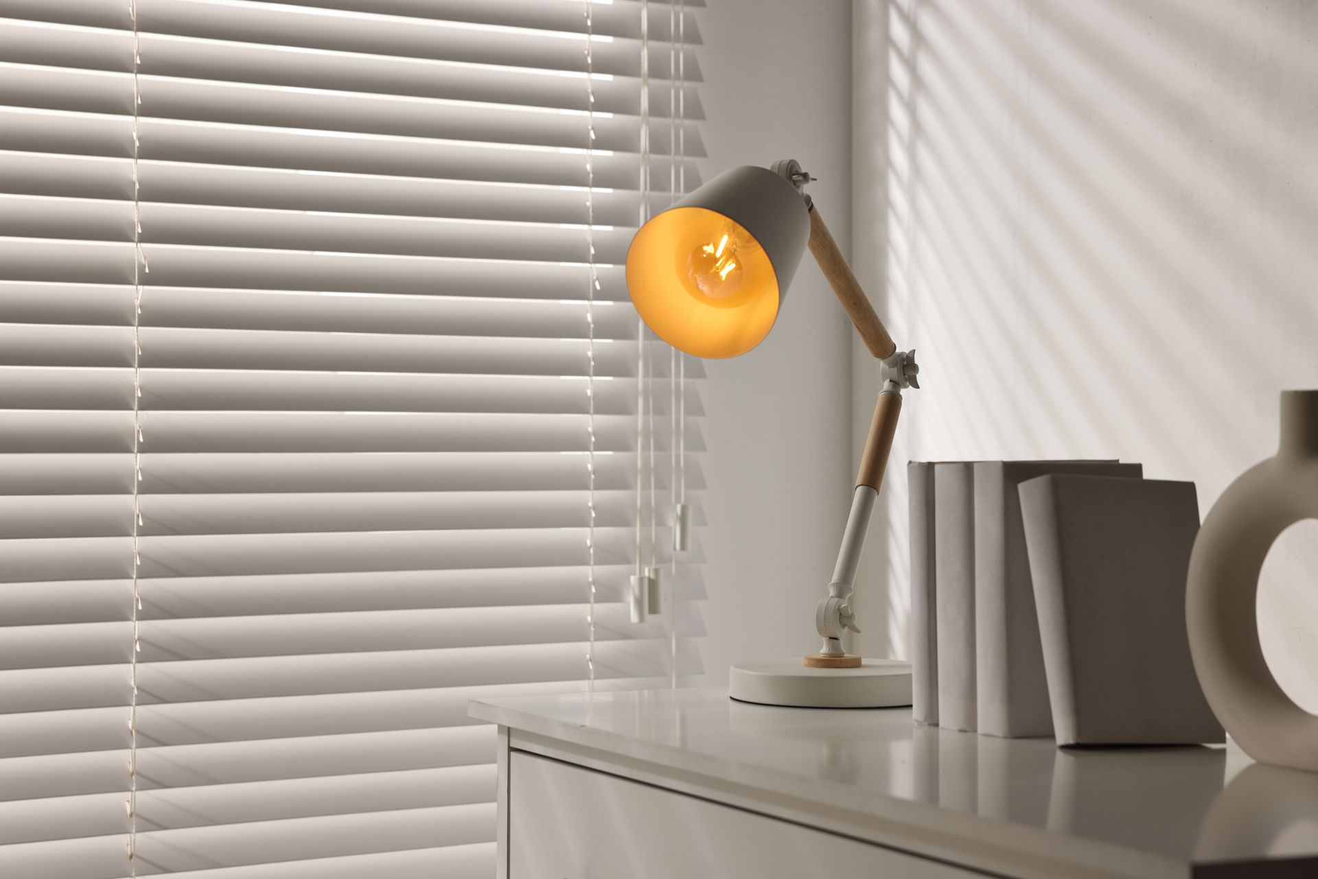 Desk lamp with warm orange light beside white blinds on a dresser