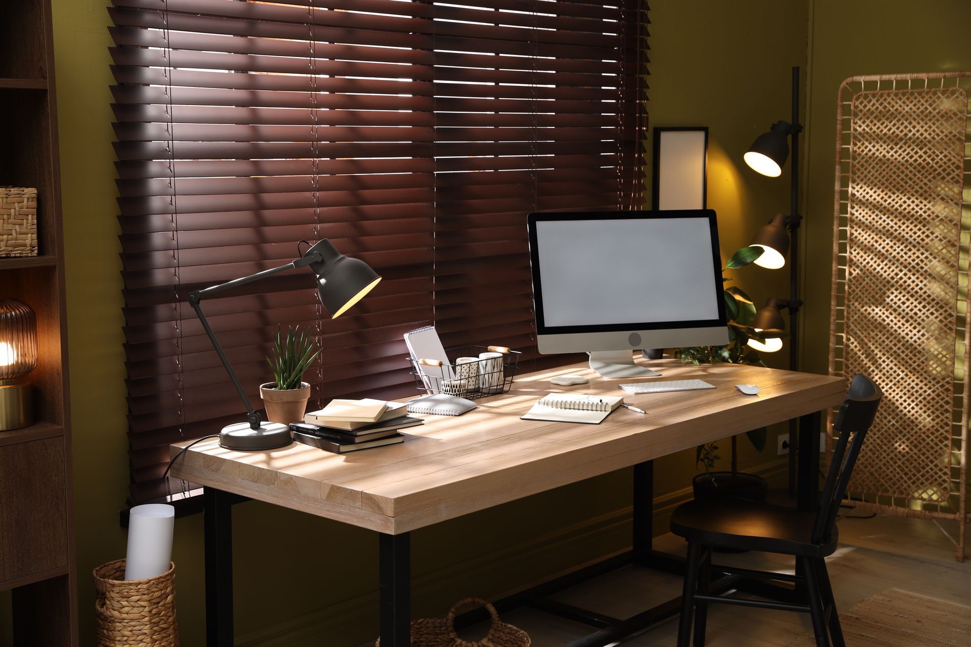 Modern home office with a wooden desk, computer monitor, lamps, papers, and window blinds.