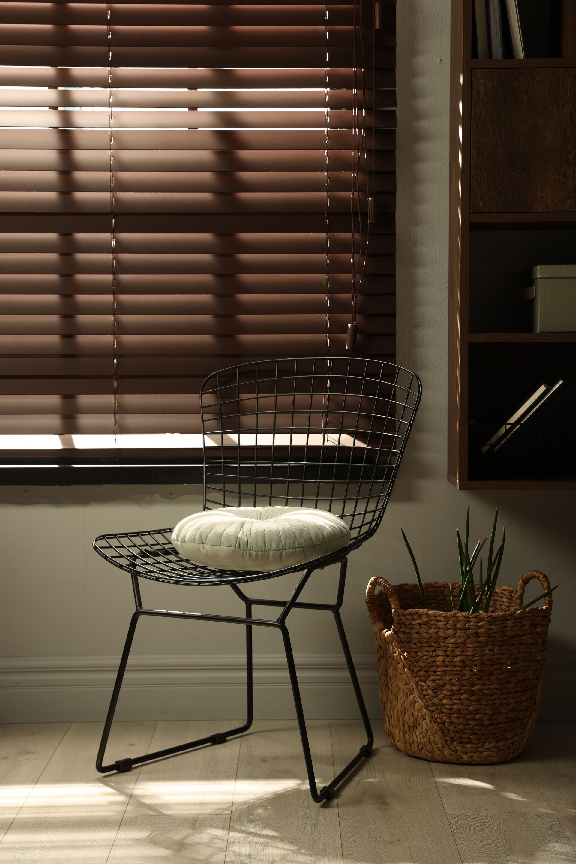 Wire chair with white cushion beside a wicker plant pot in a sunlit room with window blinds