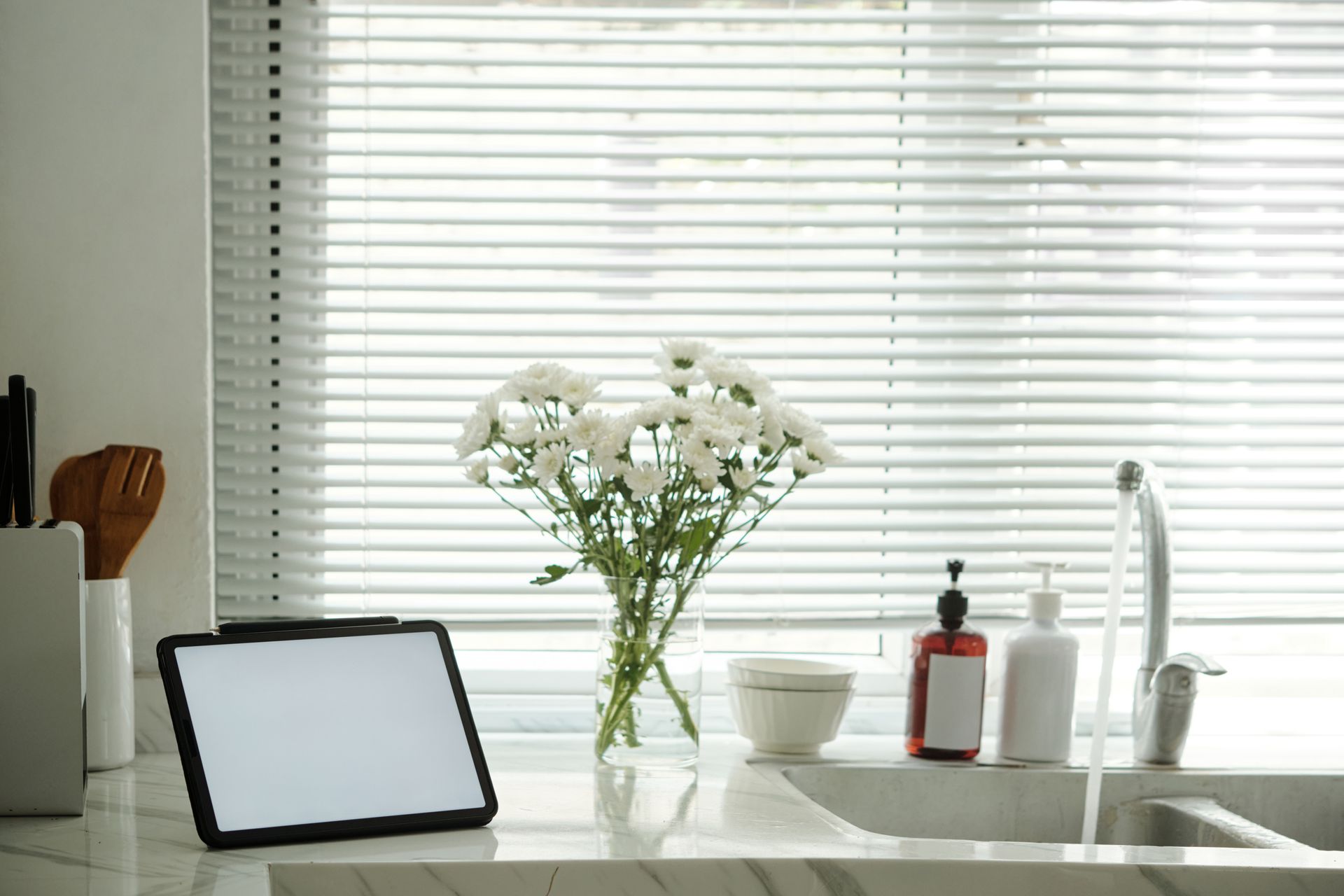 Bright kitchen sink with flowers, tablet, cup, and toiletries by a window with white blinds