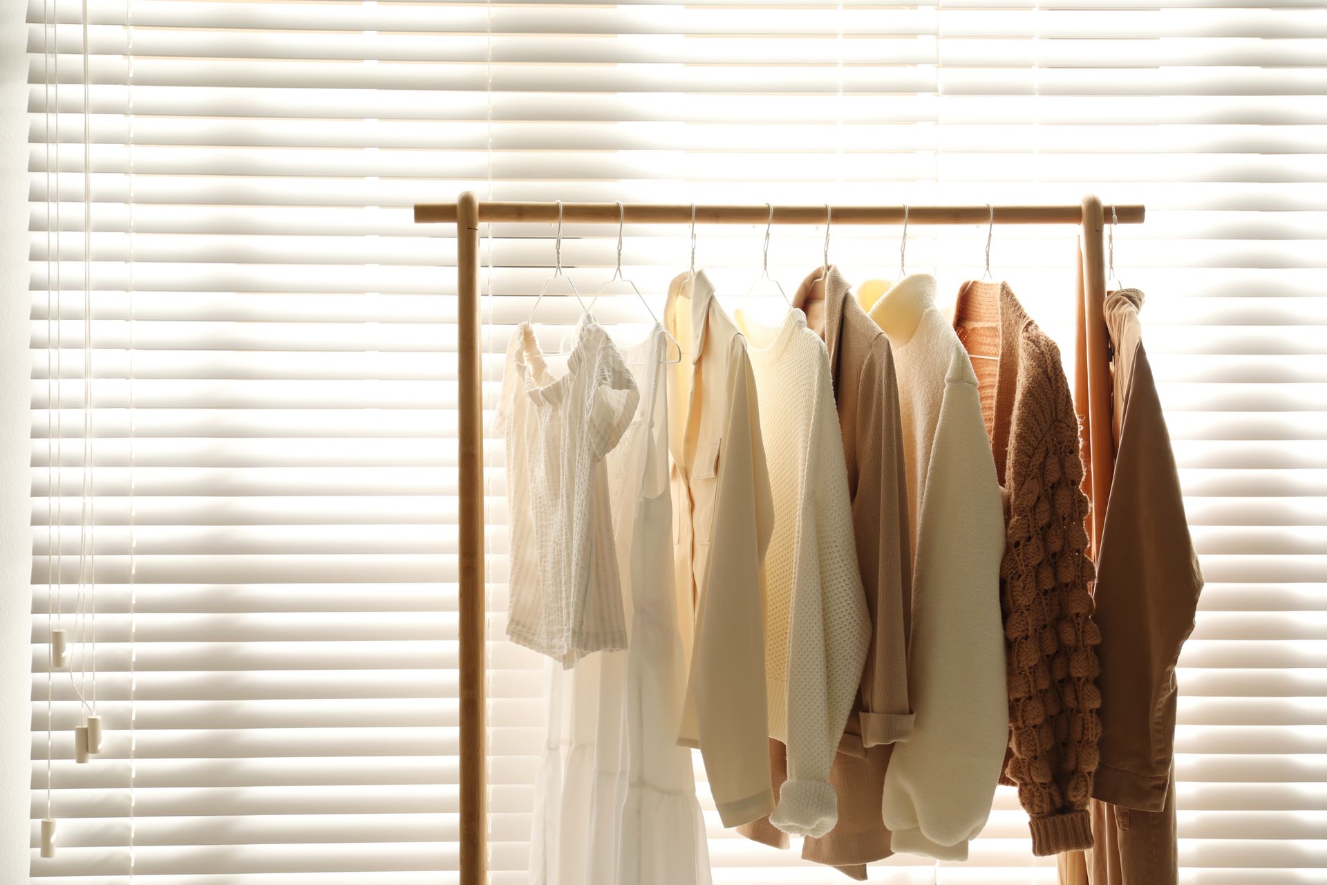 Clothing rack with neutral-toned garments hanging by a sunlit window with blinds Clothing rack with neutral-toned garments hanging by a sunlit window with blinds