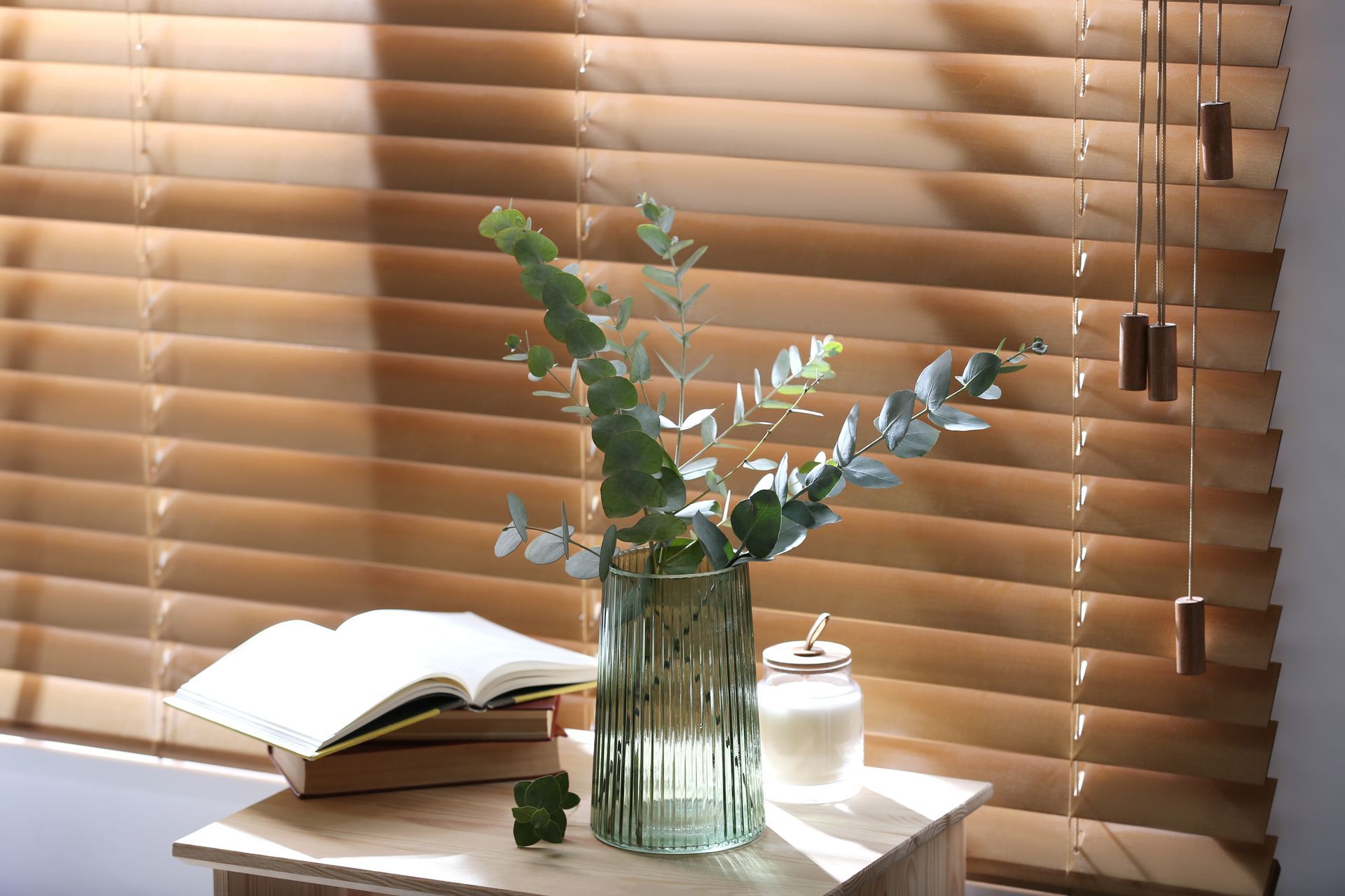 Open book, glass vase of greenery, and candle on a sunlit table by wooden blinds