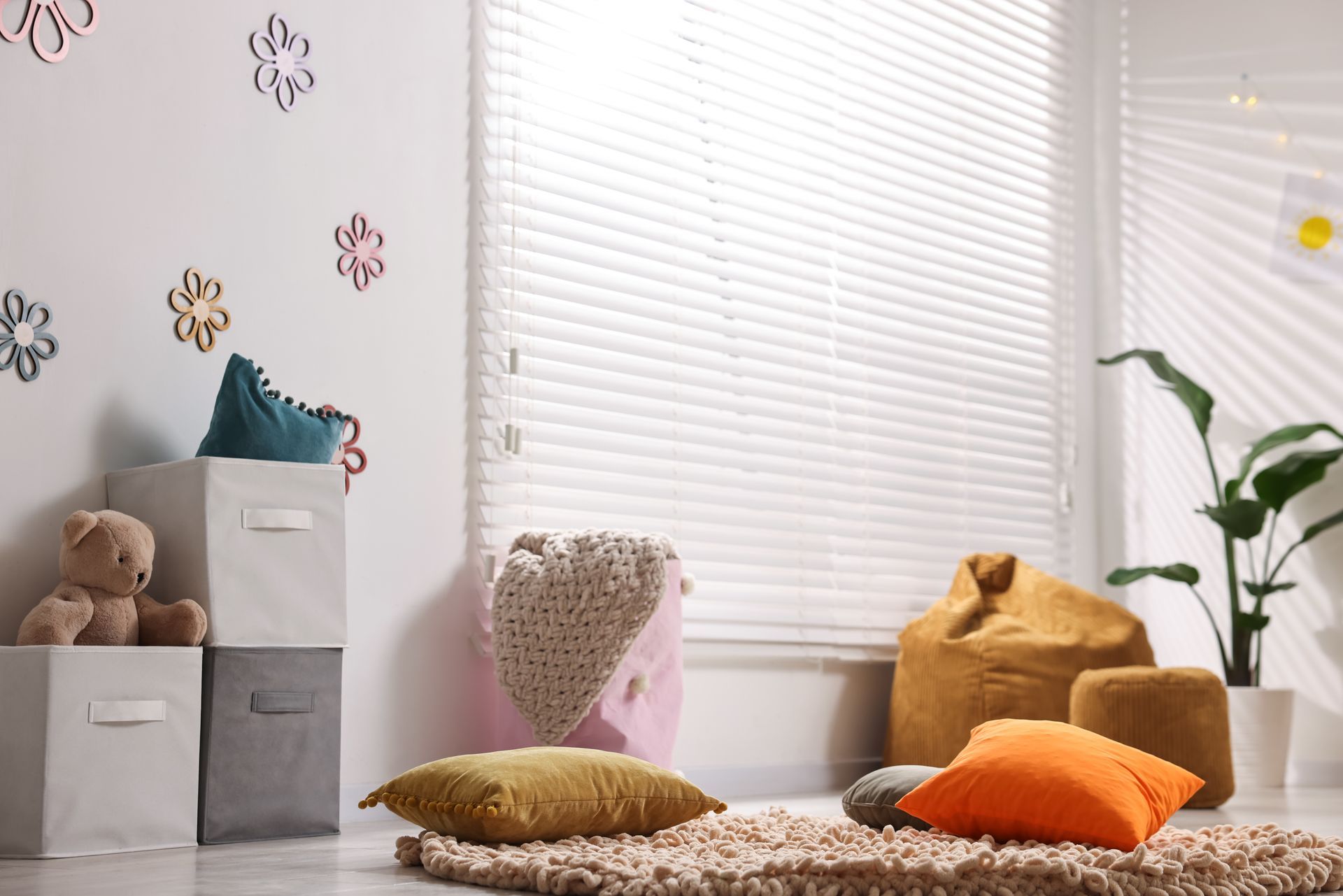 Cozy nursery corner with storage bins, plush toys, cushions, and a plant by a bright window.