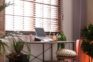 Cozy home office with a laptop on a desk, potted plants, and a cushioned chair by a window with blinds