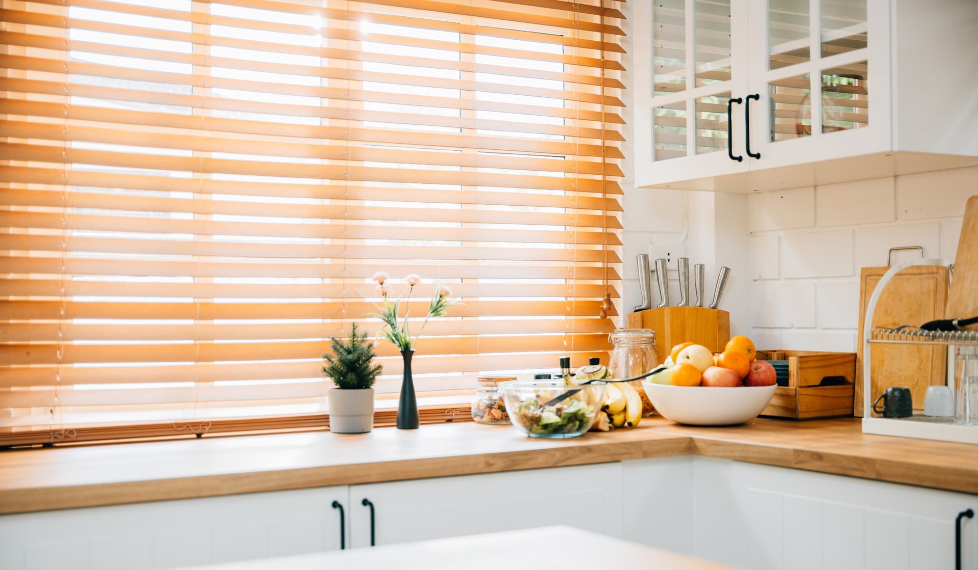 Bright kitchen windowsill with blinds, potted plants, fruit bowl, and countertop utensils