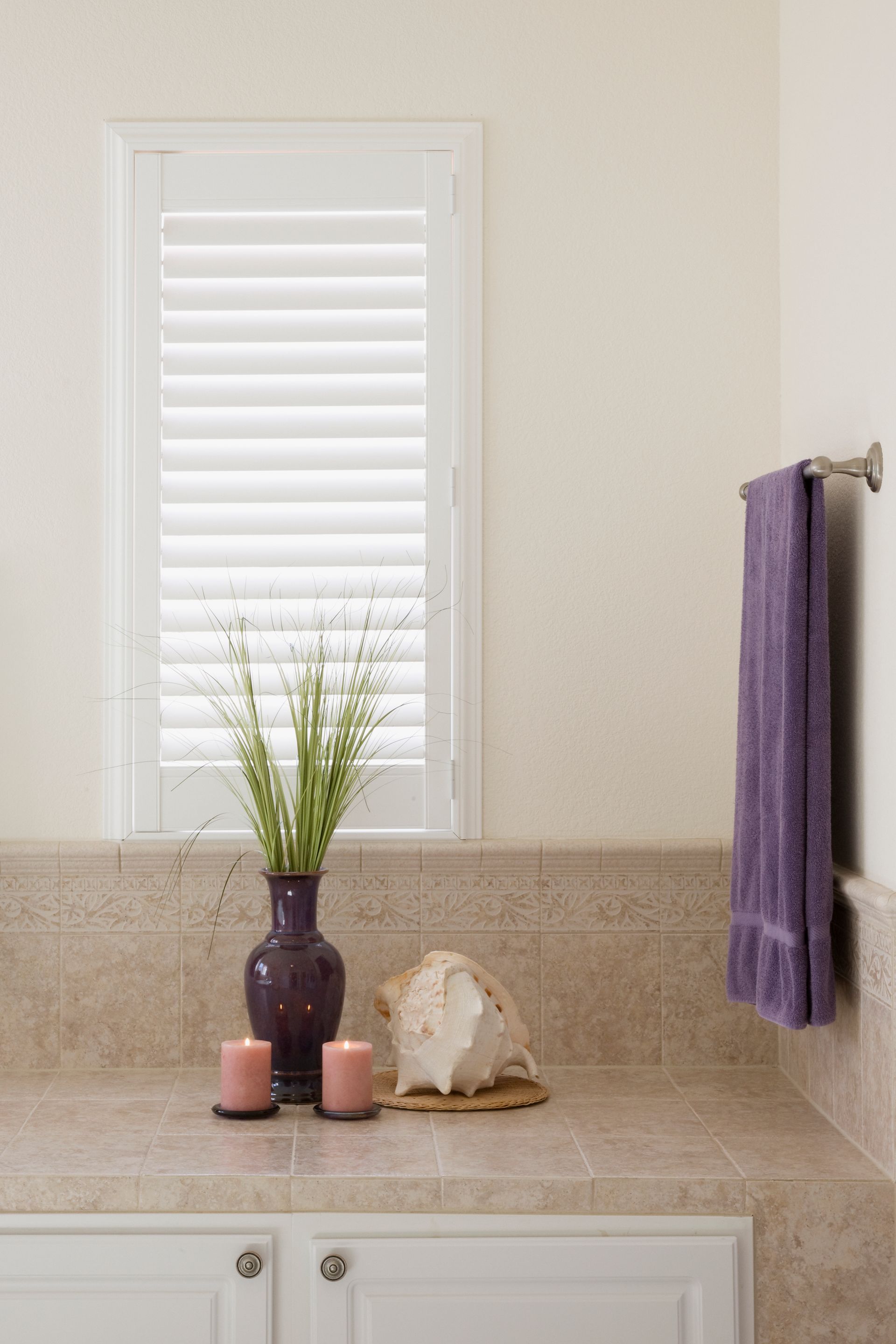 Bathroom vanity with beige tile, white shutters, purple towel, and vase of flowers on the counter Bathroom vanity with beige tile, white shutters, purple towel, and vase of flowers on the counter