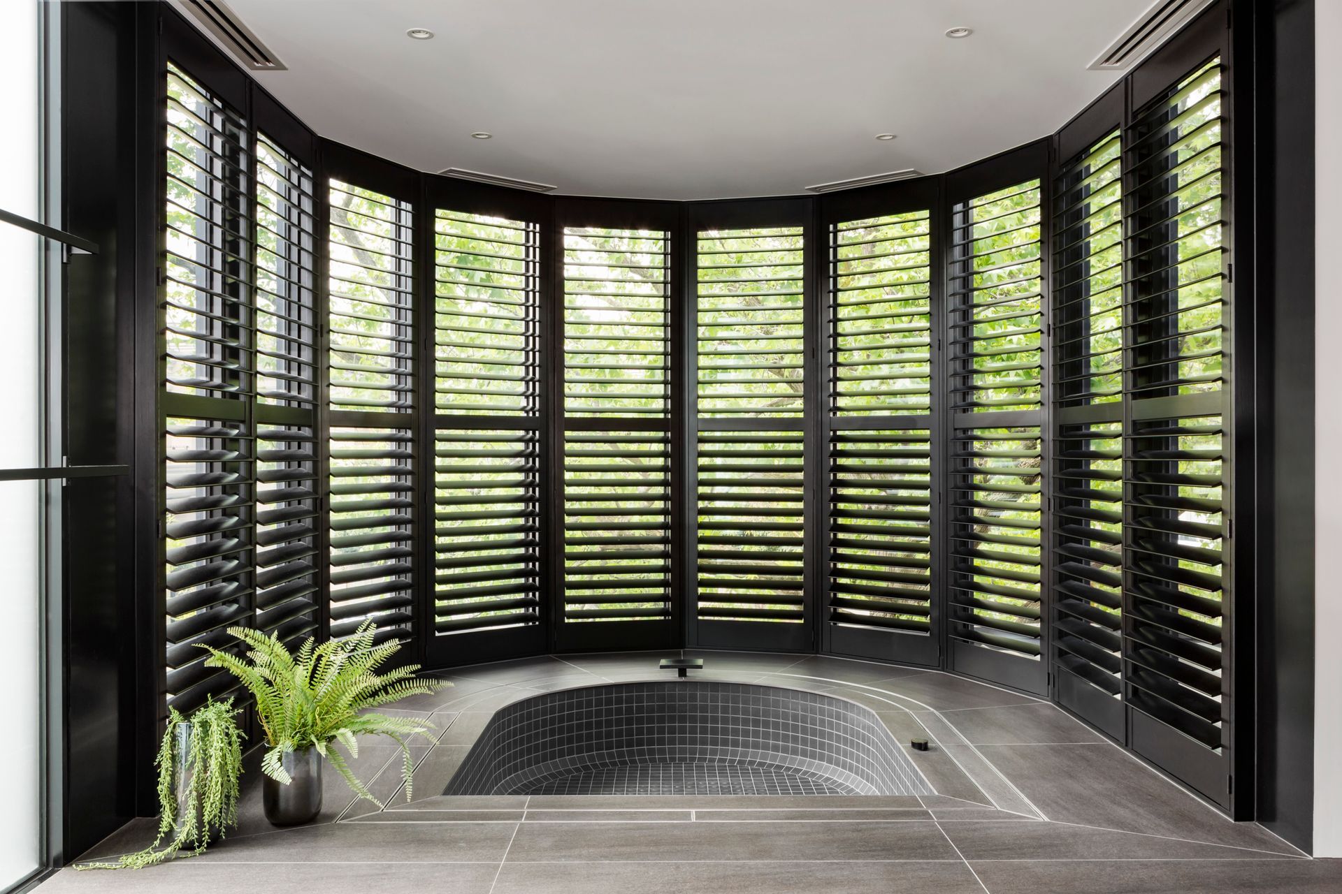 Modern bathroom with curved black shutters, a sunken tub, and potted plants Modern bathroom with curved black shutters, a sunken tub, and potted plants