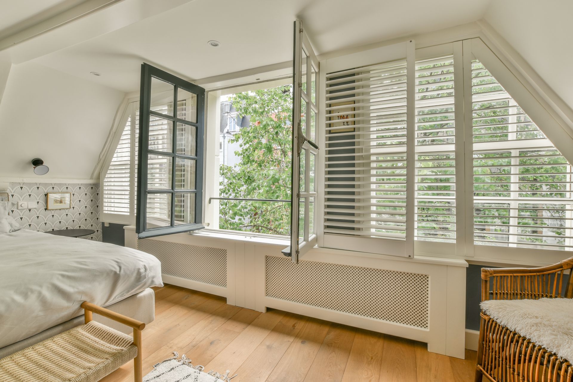 Bright bedroom with white shutters, open windows, wood floor, and a wicker chair beside a bed.