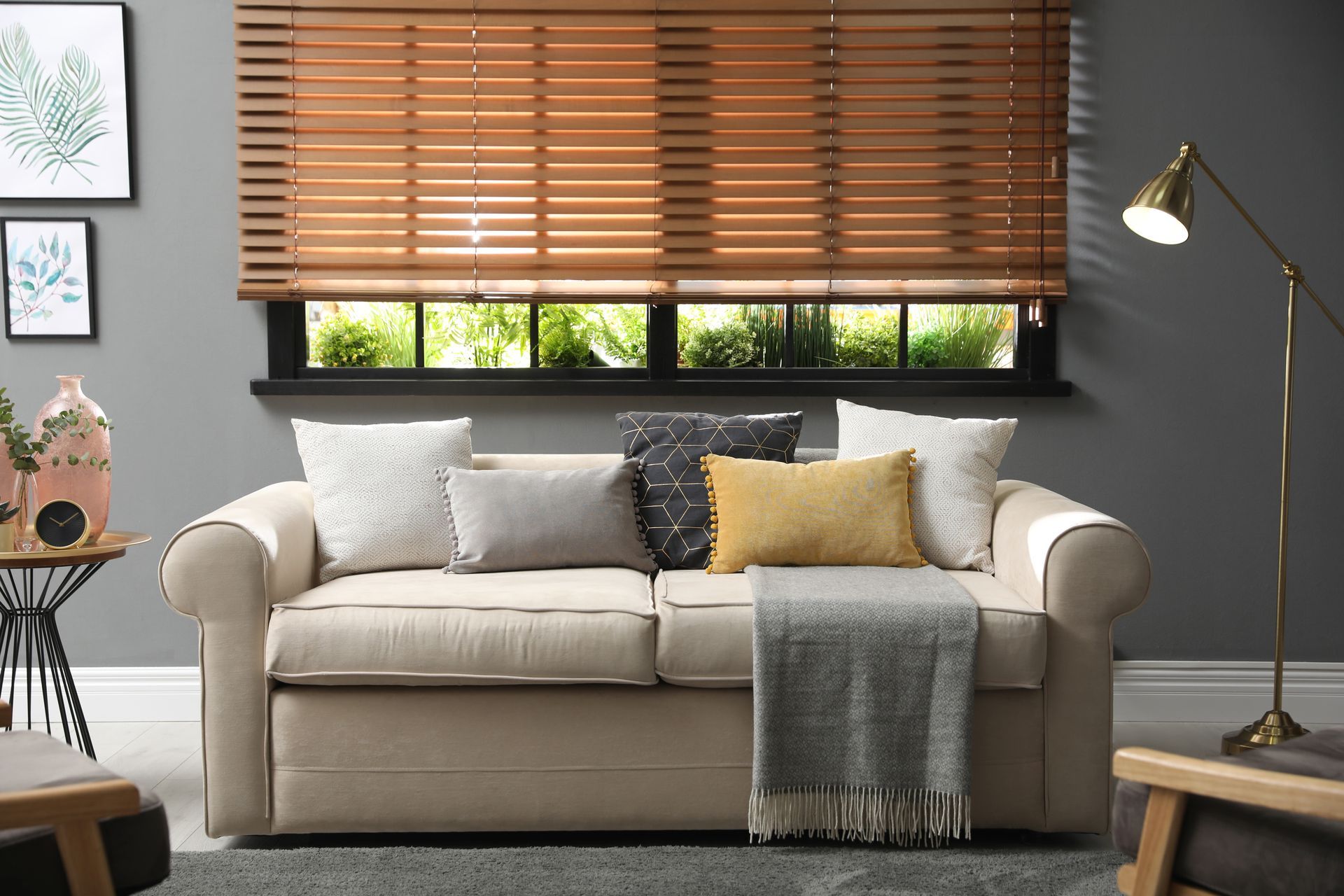 Cozy living room with beige sofa, white and yellow pillows, wooden blinds, floor lamp, and side table. Cozy living room with beige sofa, white and yellow pillows, wooden blinds, floor lamp, and side table.