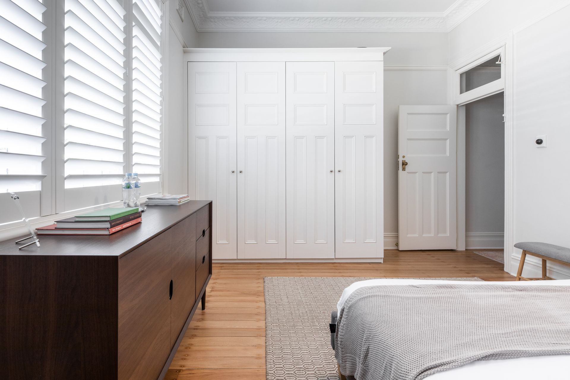 Bright bedroom with white closets, wooden dresser, gray rug, and bed near shuttered windows Bright bedroom with white closets, wooden dresser, gray rug, and bed near shuttered windows