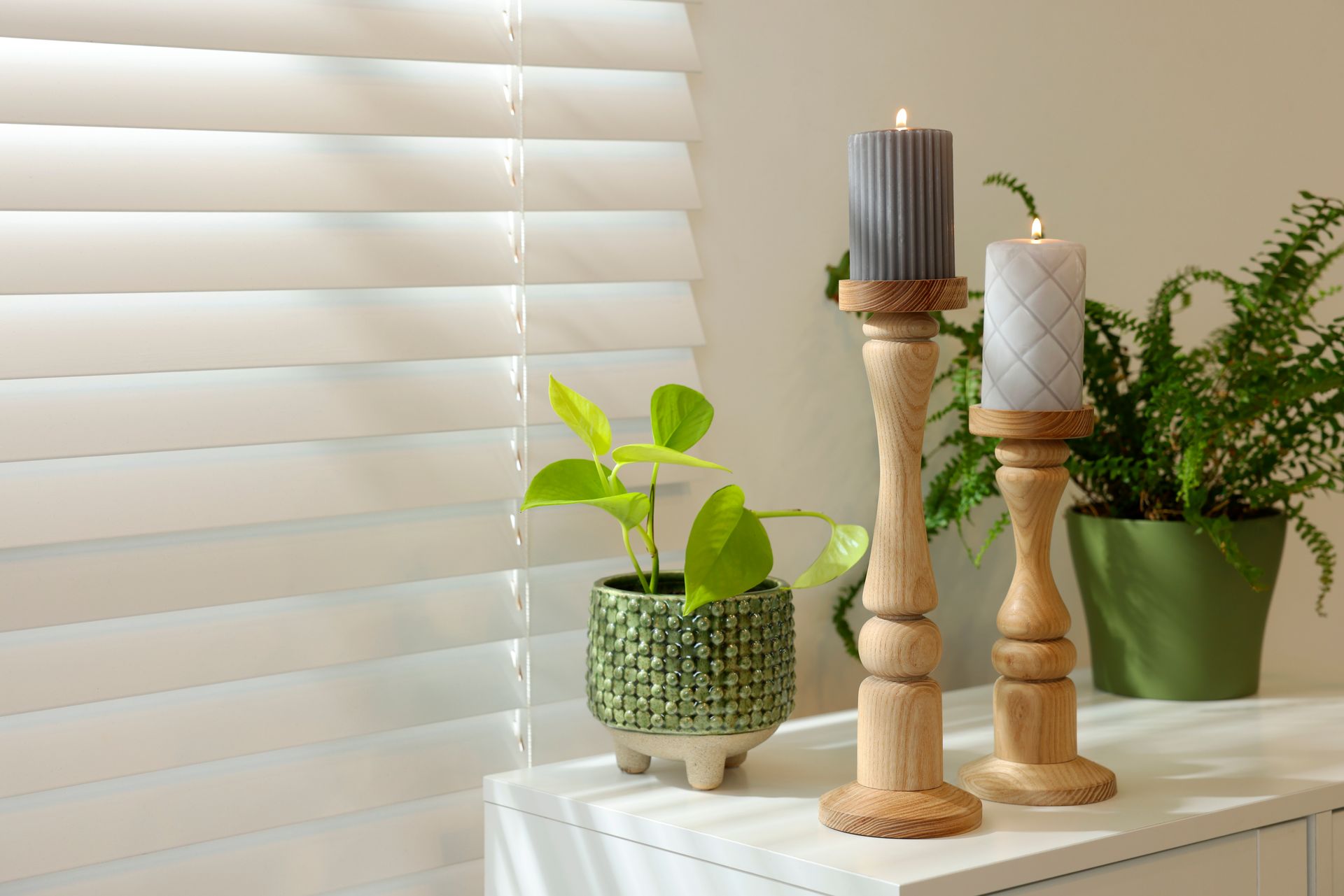Candles and potted plants on a white table by a window with blinds Candles and potted plants on a white table by a window with blinds