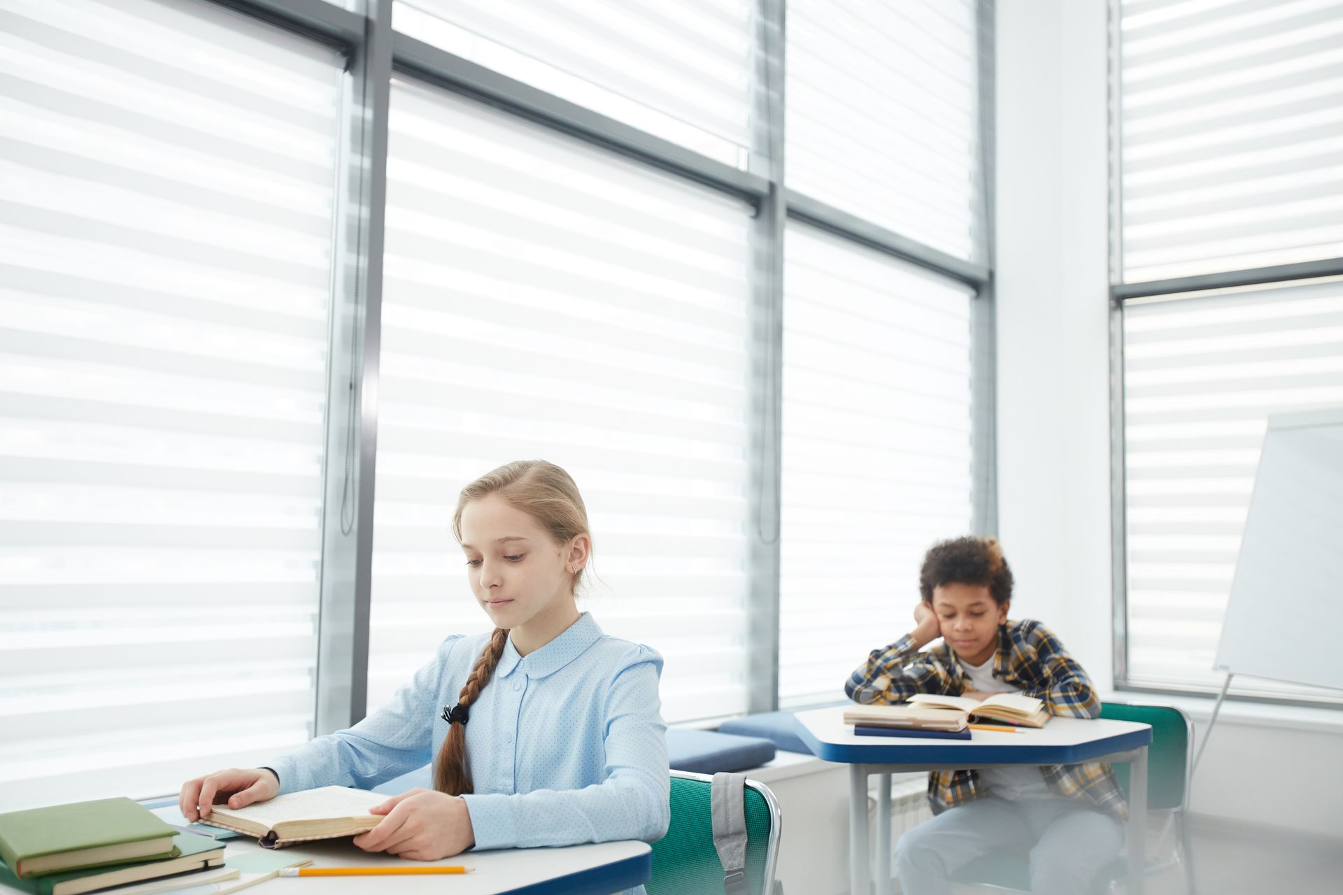 Two students working quietly at desks in a bright classroom by large windows Two students working quietly at desks in a bright classroom by large windows