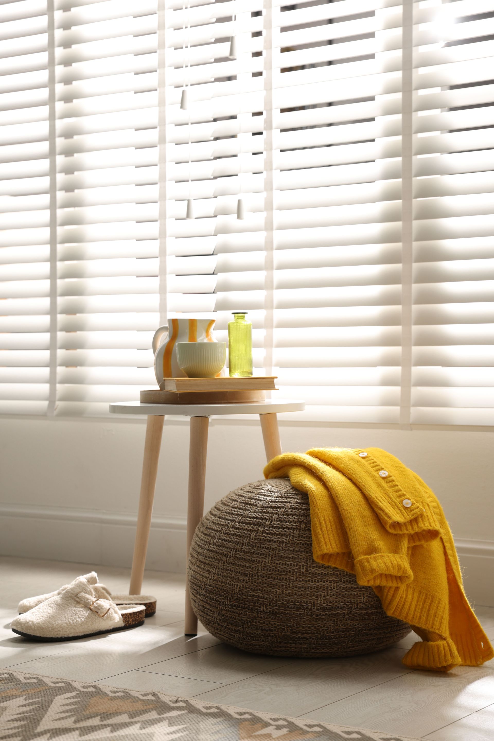 Cozy living room with a wicker pouf, yellow throw, side table, slippers, and bright window blinds Cozy living room with a wicker pouf, yellow throw, side table, slippers, and bright window blinds